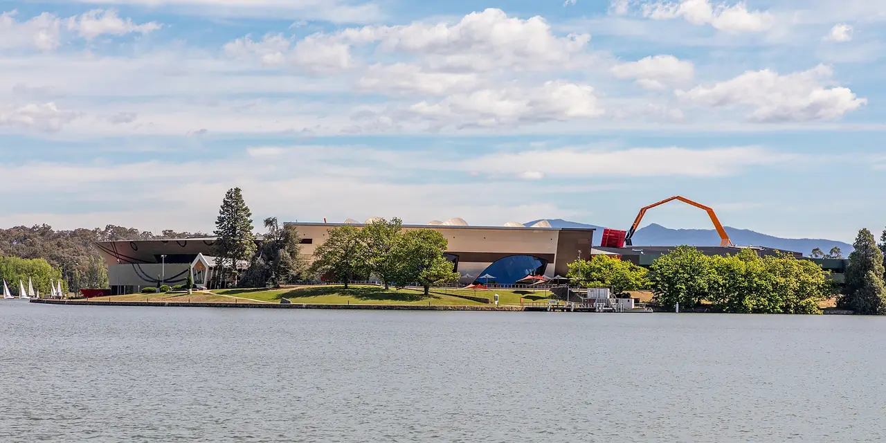 พิพิธภัณฑ์แห่งชาติออสเตรเลียมองข้ามทะเลสาบ Burley Griffin มีโครงสร้างสีส้มยื่นออกมาด้านหลัง ภาพจาก Wikimedia Commons ผู้ถ่าย Dietmar Rabich สัญญา CC BY-SA 4.0 ถ่ายปี 2019