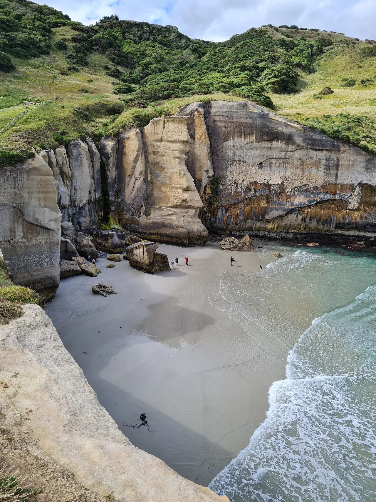 หาดเล็กใต้หน้าผาหินปูนที่ Tunnel Beach มีคนยืนอยู่บนทรายและคลื่นเข้าชายฝั่ง ภาพจาก Wikimedia Commons ผู้ถ่าย ChristchurchHistorian สัญญา CC BY-SA 4.0 ไม่ระบุปีที่ถ่าย