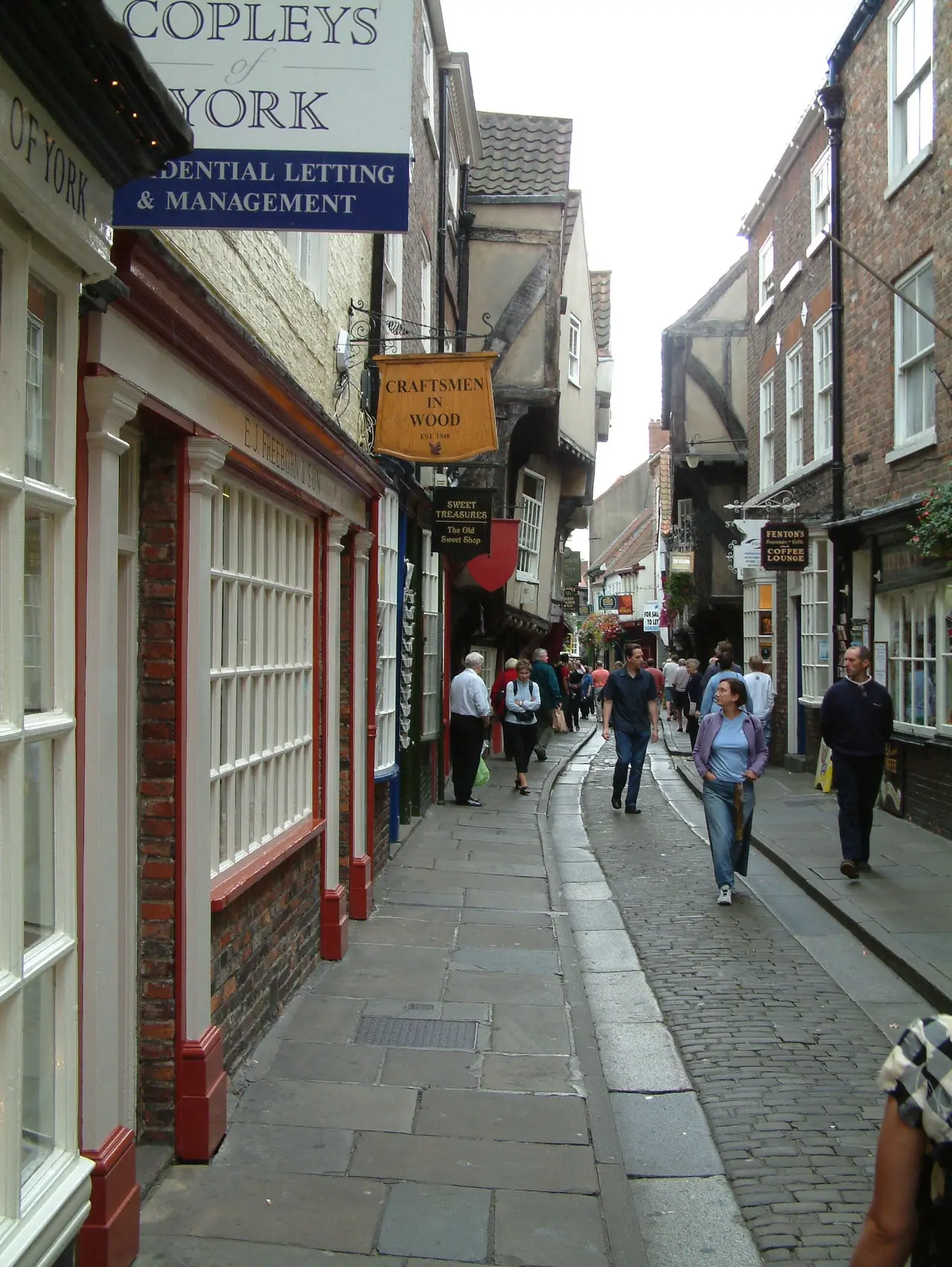ถนน The Shambles ในยอร์ก มีบ้านไม้เก่าและทางเดินแคบ ภาพจาก Wikimedia Commons ผู้ถ่าย Malcolm Neal สัญญา CC BY-SA 2.0 ไม่ระบุปีที่ถ่าย ร้านค้าในภาพอาจเปลี่ยนไปแล้ว