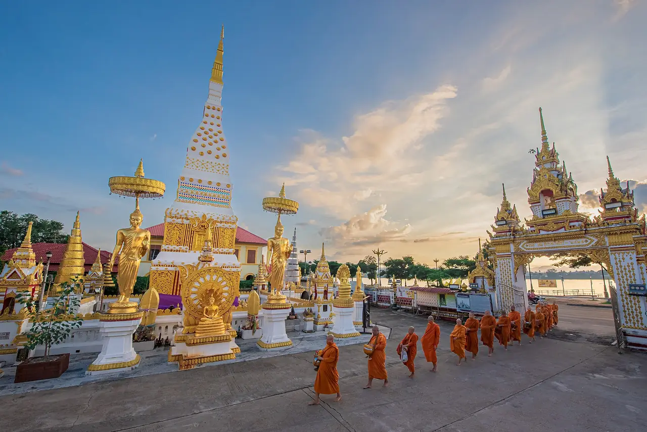 พระธาตุนคร วัดมหาธาตุ นครพนม ยามเย็น พระสงฆ์เดินเรียงแถวหน้าองค์พระธาตุ และริมโขงอยู่ด้านขวาของภาพ ภาพจาก Wikimedia Commons ผู้ถ่าย JayaJoe สัญญา CC BY-SA 4.0 ตามชื่อไฟล์เป็นวัดมหาธาตุ นครพนม ไม่ระบุวันที่ถ่าย