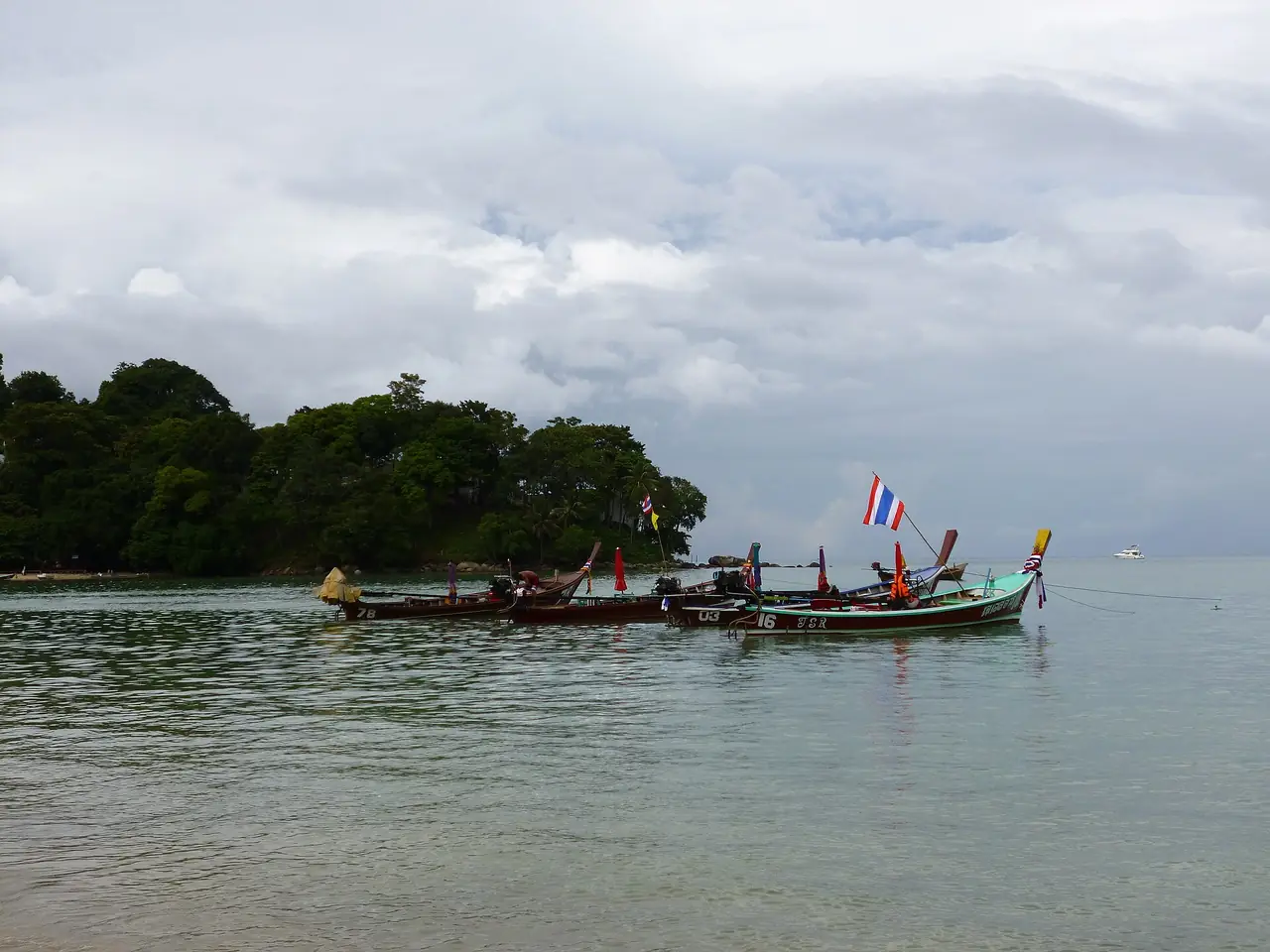 เรือหางยาวจอดนิ่งในอ่าวที่มีธงชาติไทยโบกบนเรือและป่าเขียวด้านหลัง ฟ้าครึ้ม ภาพจาก Wikimedia Commons ผู้ถ่าย Arnaud-Victor Monteux สัญญา CC BY-SA 4.0 ตามคำบรรยายไฟล์ถ่ายที่หาดป่าตอง พ.ย. 2555 อาจไม่ใช่หน้าตาปัจจุบัน