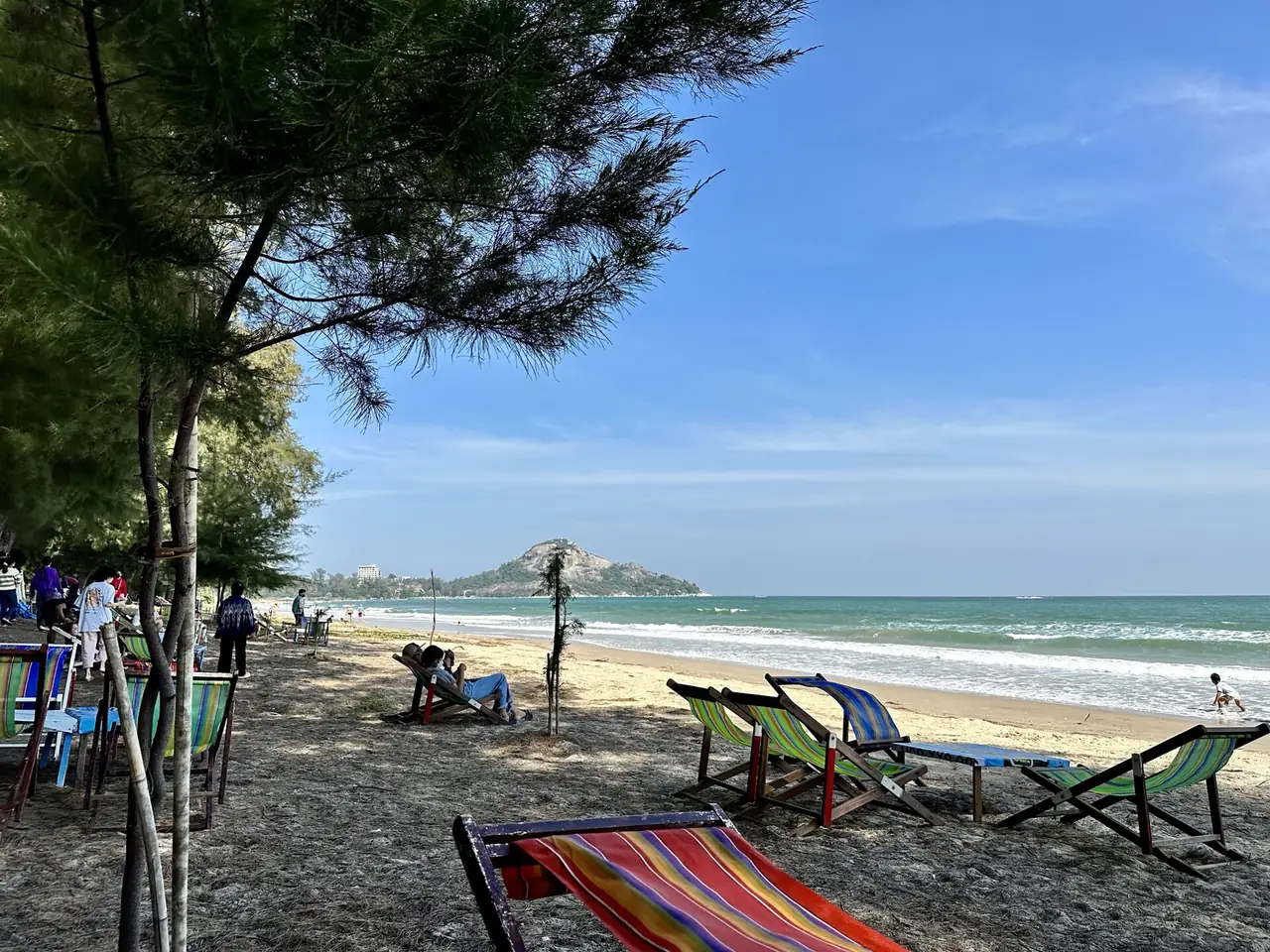 เก้าอี้ผ้าลายสีสดวางใต้ต้นสนริมหาด ทะเลสีเขียวและเนินเขาเล็ก ๆ อยู่ไกลทางขวา มีคนนั่งพักและเด็กวิ่งเล่น ภาพจาก Wikimedia Commons ผู้ถ่าย Slyronit สัญญา CC BY-SA 4.0 ชื่อไฟล์ว่า Suan Son Pradiphat Beach ไม่ระบุปีที่ถ่าย เก้าอี้และร้านอาจเปลี่ยนไปแล้ว