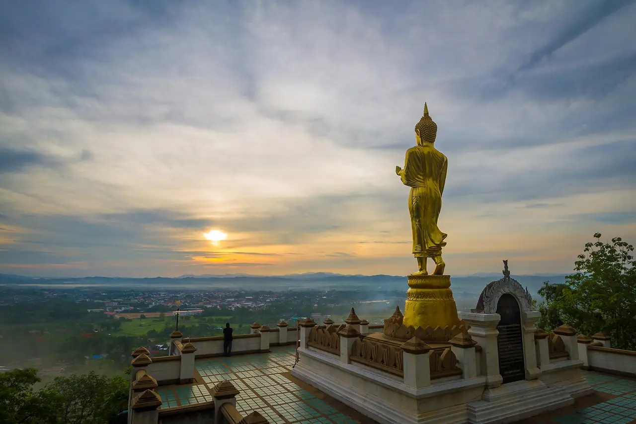 พระพุทธรูปสีทองยืนบนฐานที่ลานชมวิวของวัดพระธาตุเขาน้อย พระอาทิตย์ดวงเล็กอยู่ต่ำเหนือขอบฟ้า เมืองและทุ่งอยู่เบื้องล่างและมีคนยืนมองอยู่ที่ขอบลาน ภาพจาก Wikimedia Commons ผู้ถ่าย Manoonp สัญญา CC BY-SA 4.0 ไม่ลงวันที่ถ่ายในรายละเอียดที่เราเปิดดู เป็นภาพประกอบบรรยากาศช่วงแสงต่ำ