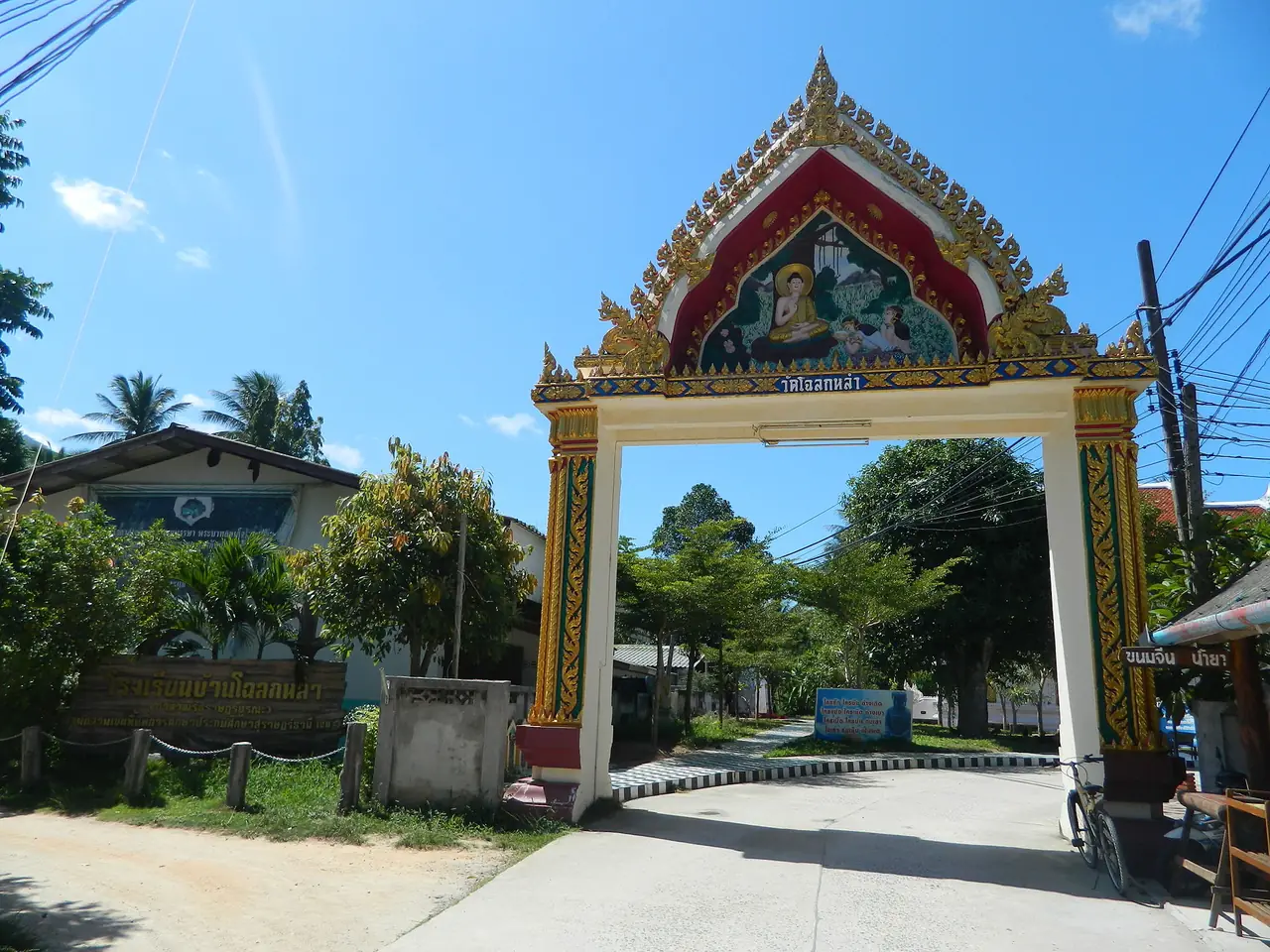 ซุ้มประตูทางเข้าวัดโฉลกหลำ มีภาพพุทธประวัติบนหน้าจั่วสีแดงทอง และถนนคอนกรีตเข้าหมู่บ้าน โฉลกหลำคืออ่าวทางเหนือที่หน้าเว็บ Wonderland ใช้บอกระยะ ราว 4 กม. ภาพจาก Wikimedia Commons ผู้ถ่าย Леонид Каратасков สัญญา CC BY-SA 3.0
